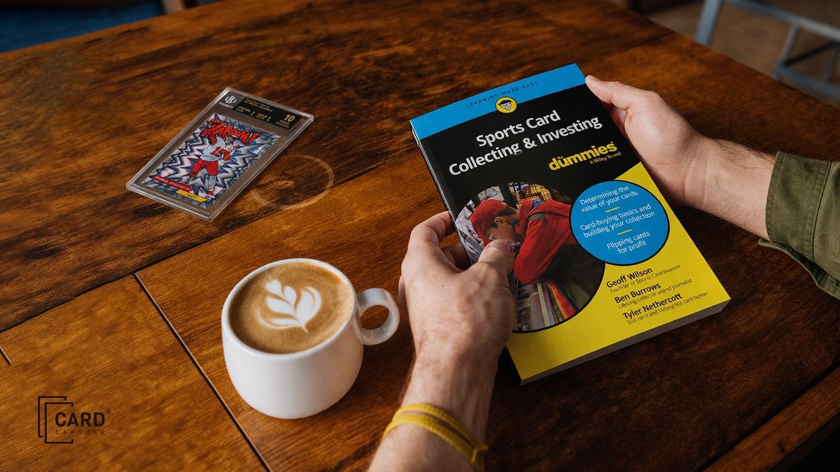 Hands holding a sports card collecting guidebook on a wooden table beside a latte and a graded card